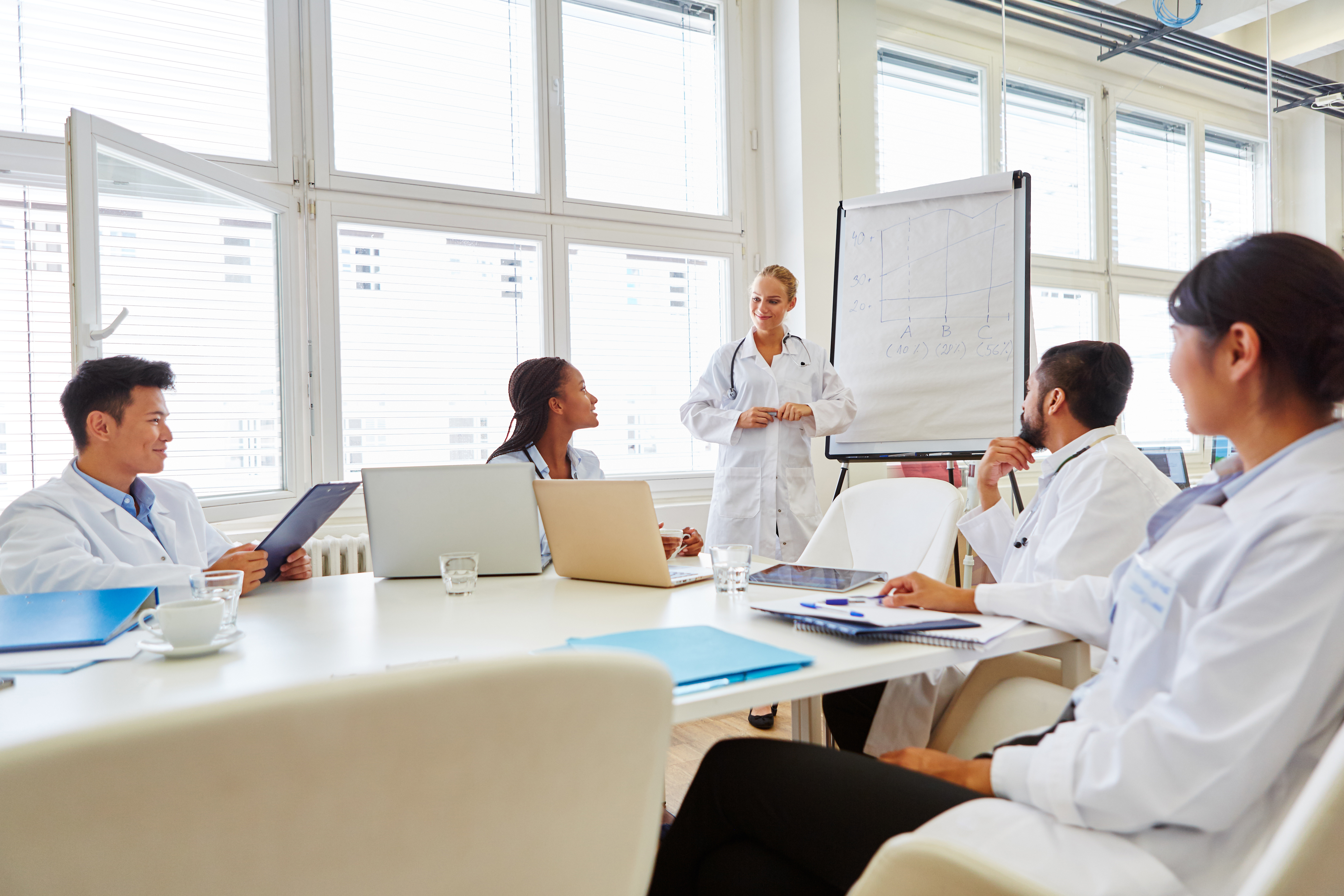 Group of health care providers sitting around a table looking at another health care provider standing in front of a white board.