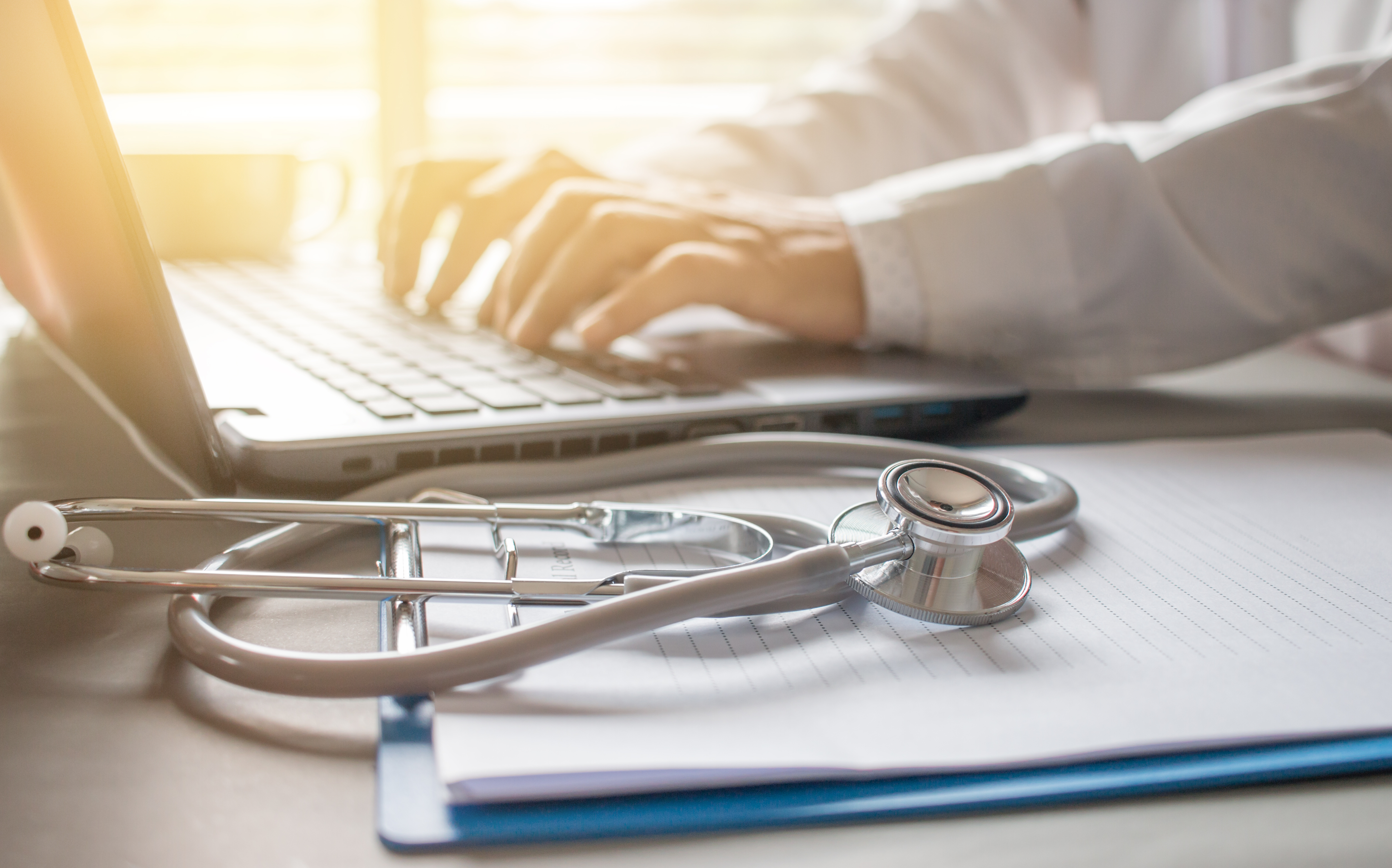 Image of a health care provider's hands hovering over a laptop computer beside a stethoscope laying on top of a medical file.