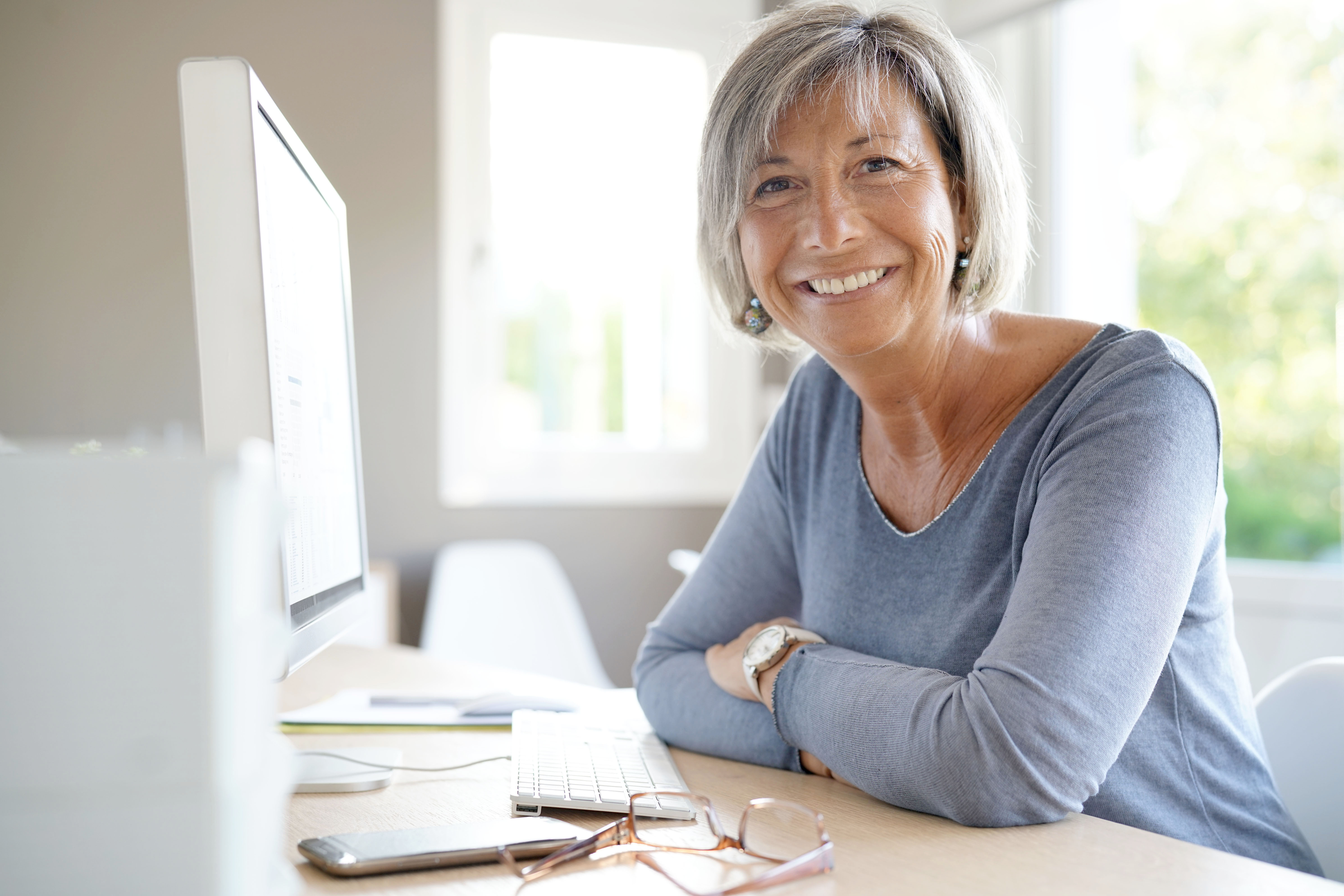 Image of older adult woman sitting at a computer smiling and facing the camera.