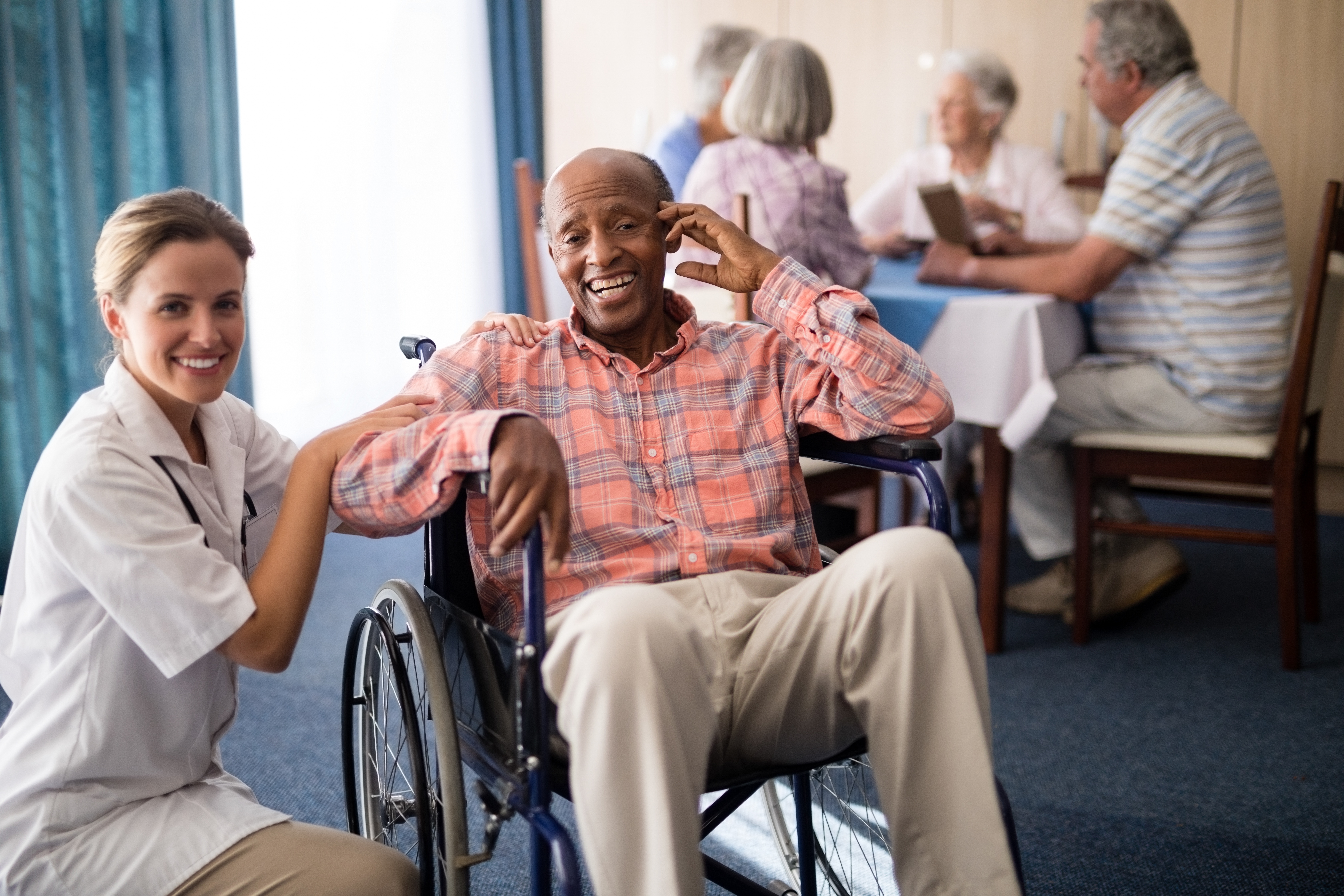 Image of an older man smiling while sitting in a wheelchair while a nurse kneels down beside him and smiles toward the viewer.