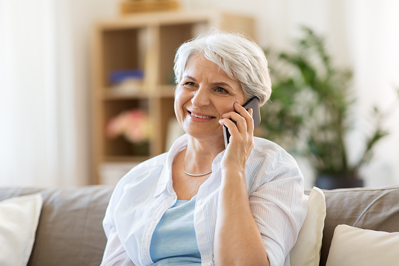 Image of older woman sitting on a couch and talking on a phone she is holding with her left hand.