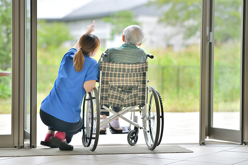 Image of two people, one in a wheel chair and one kneeling beside the other, looking out double doors.