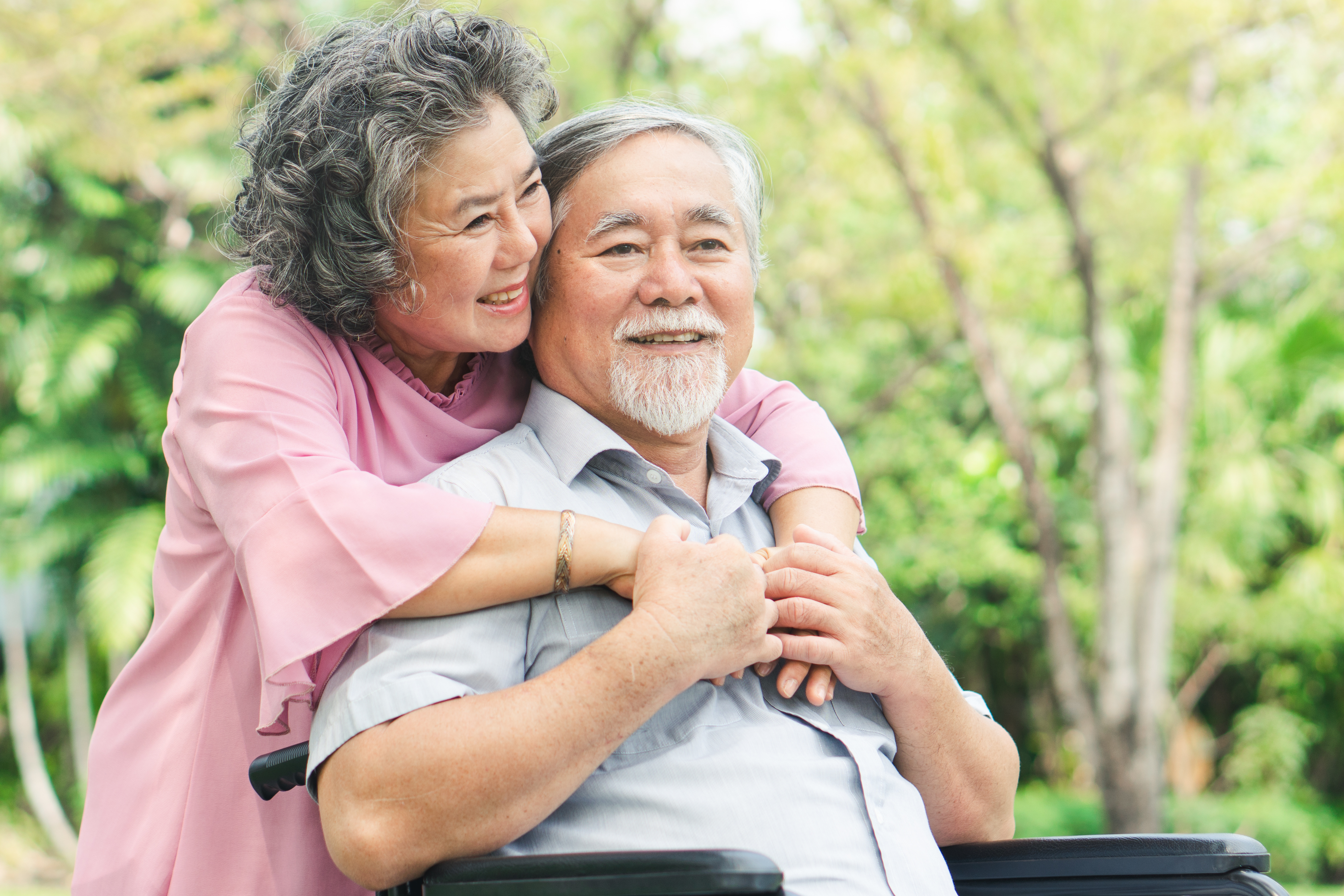 Image of older Asian woman putting her arms around an older Asian man who is sitting in a wheel chair.