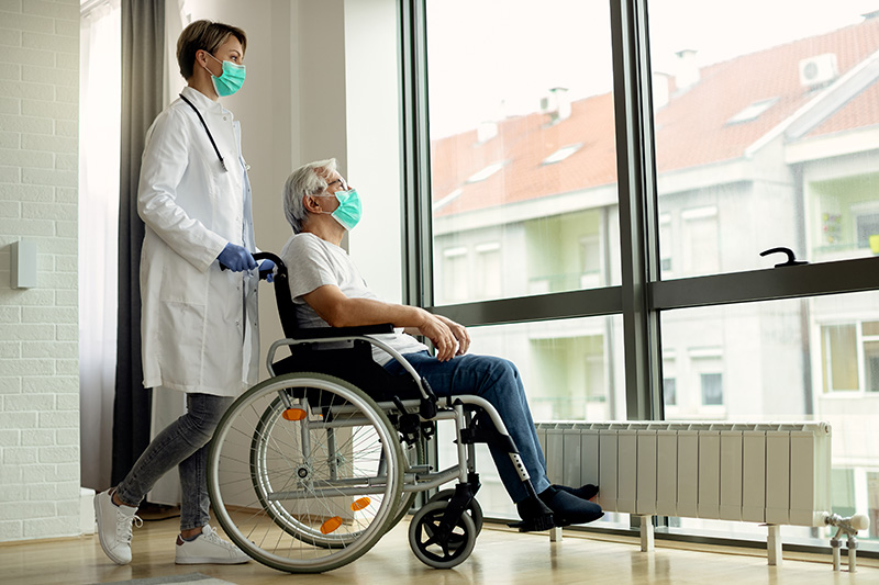 Image of older man in a wheelchair wearing a green mask and looking out the window while a medical professional wearing a white coat and green face mask stands behind the wheelchair with his hands on the chair.