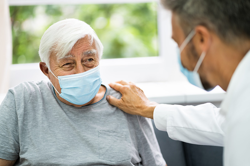 Image of a male doctor visible from the side wearing a white coat and surgical mask, with his right hand on the left shoulder of a seated, older white man with white hair wearing a gray shirt and light blue surgical mask.