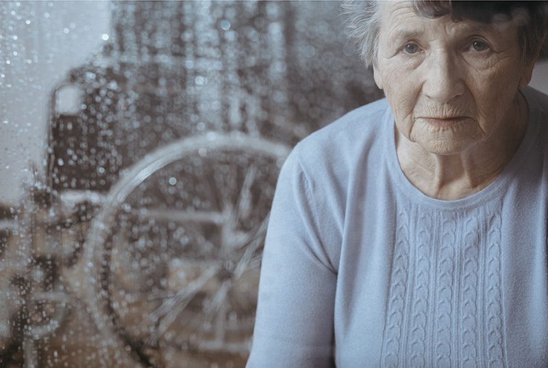 Image of older woman looking sad with a rain covered window behind her through which you can see a wheelchair