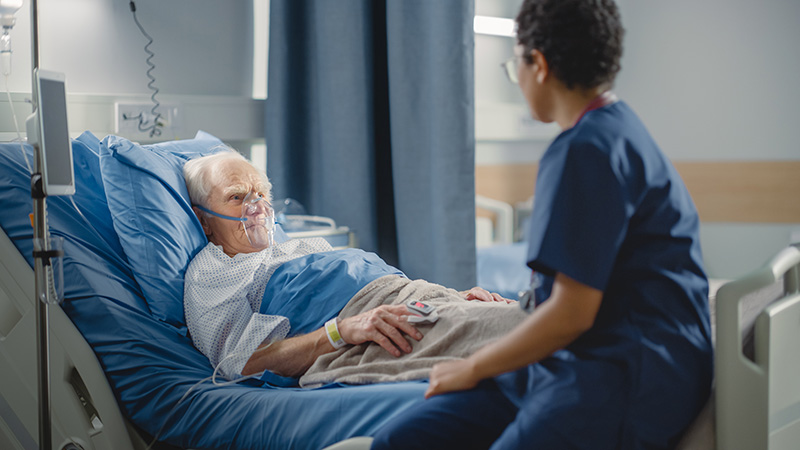 Image of a nurse wearing navy scrubs sitting at the bedside of an older man wearing an oxygen mask