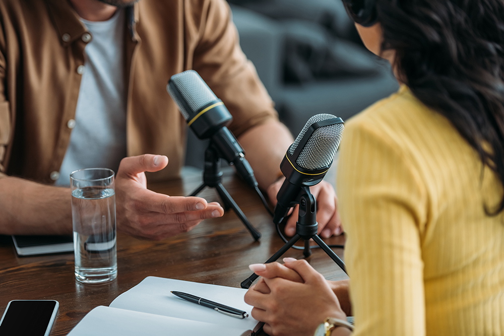 Image of two people talking on a microphone between them as if recording a podcast.