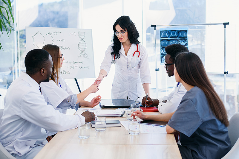 Image of medical staff sitting around a table while looking at another medical professional standing at a white board.