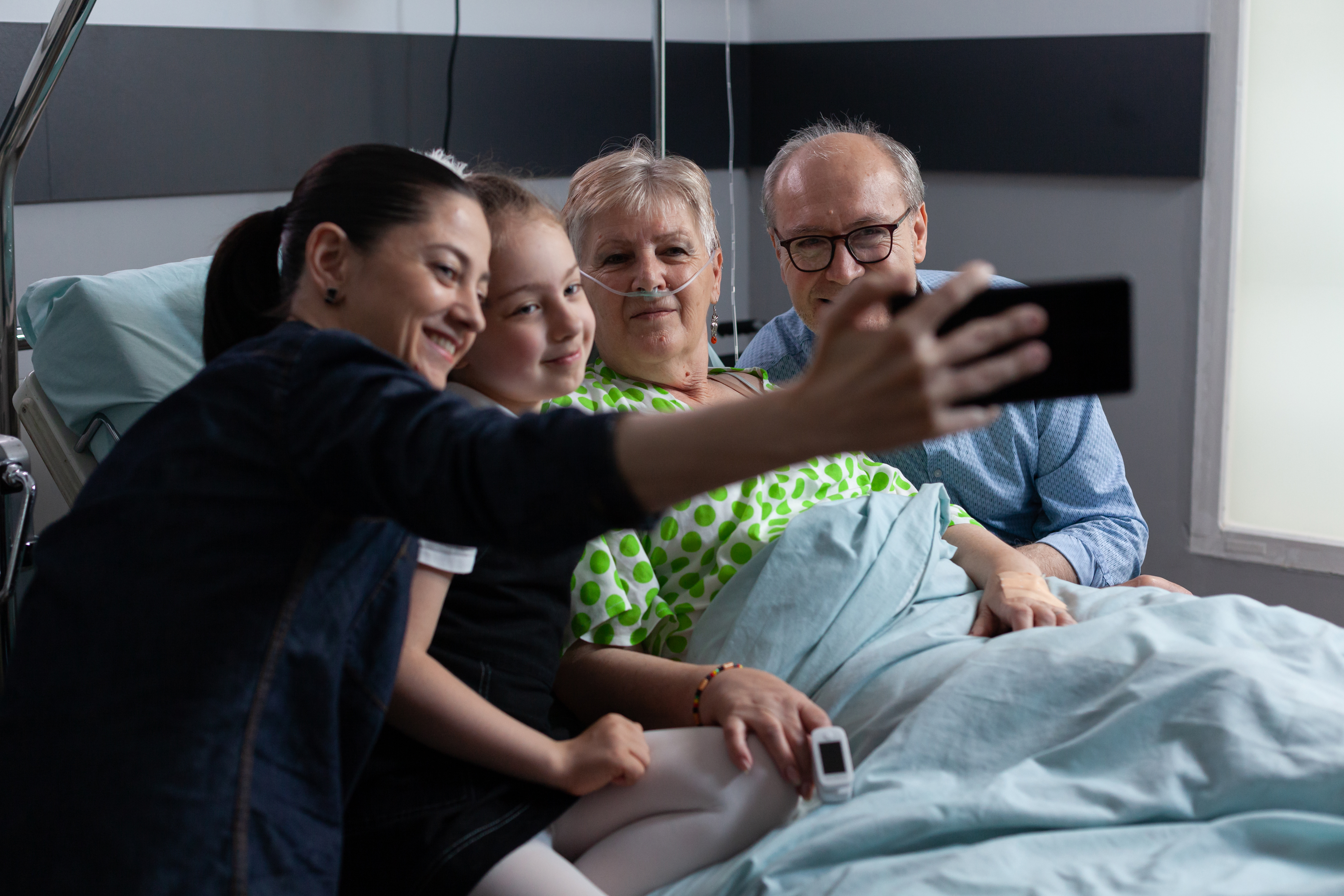 Elderly patient smiling for selfie alongside husband, daughter, granddaughter in older people medical clinic observation room.