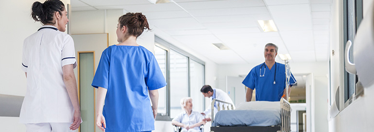 Image of hospital hallway with a man on the right pushing a stretcher and two nurses walking together in the opposite direction on the left.