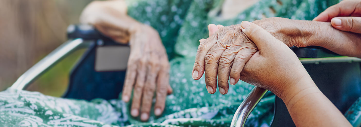 Image of older African American woman wearing a green dress and sitting in a wheel chair and whose left hand and forearm are held by another African American person.