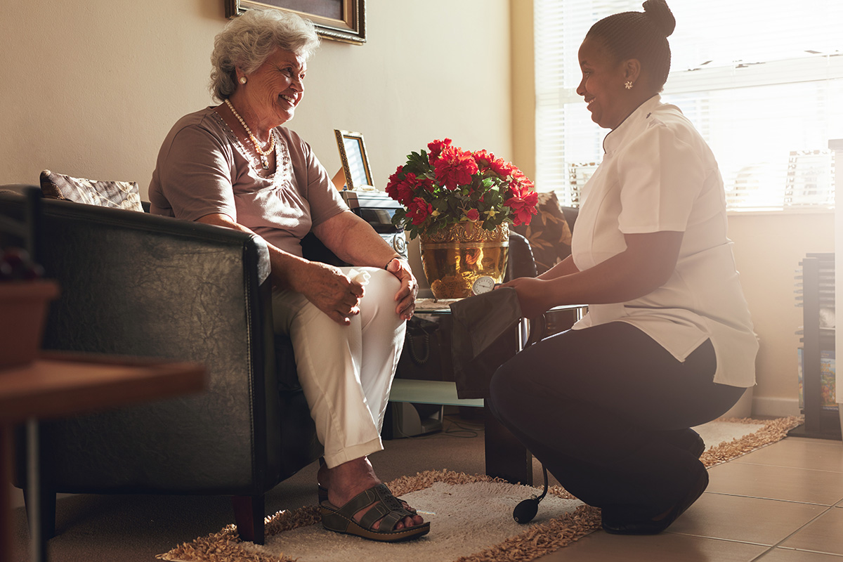Female nurse kneeling down in front of smiling senior woman at nursing home