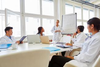 Image of health care providers sitting around a table while another provider speaks in front of a whiteboard.
