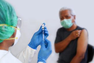Image of African-American woman wearing a green mask lifting up the left sleeve of her shirt as a health care worker wearing a green hair net, white mask and blue medical gloves prepares a syringe.