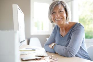 Image of older woman sitting in front of a white computer, turned toward the camera and smiling