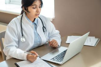 Female doctor wearing a Bluetooth ear piece and stethoscope around her neck sitting at a lap top computer having a video call.