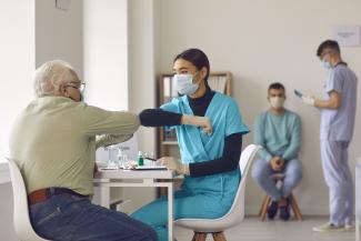 Young nurse in face mask bumping elbows with senior man before giving him vaccine