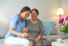Smiling nurse in blue uniform hugs old lady while sitting on grey sofa in nursing home. 