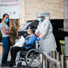 Healthcare professional escorting patient in wheelchair, while patient interacts with woman with mask on.