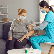 Safe vaccination for old people. Mature woman in medical face mask getting flu or Covid-19 vaccine sitting on sofa at home.