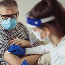 Young woman nurse with surgical mask and face shield giving injection to senior man at home or in a nursing home. 