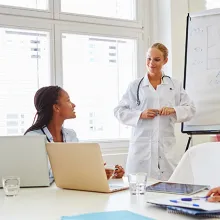 Healthcare providers in white coats sitting around table listening to doctor present.