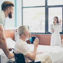 Male pushing elderly man in wheelchair out of room waving to clinician