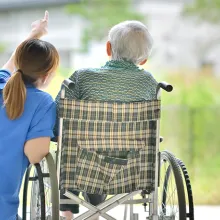 Female healthcare worker kneeling beside an older male in a wheelchair looking out the window on a nice day.