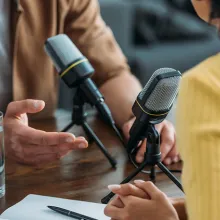 Image of two people talking on a microphone between them as if recording a podcast.