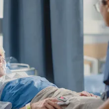 Friendly Head Nurse Connects Finger Heart Rate Monitor or Pulse Oximeter to Elderly Patient Wearing Oxygen Mask Resting in Bed.