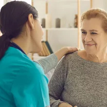 Friendly doctor supporting senior woman. Happy young nurse or clinician and her older adult patient sitting on sofa, looking at each other, holding hands, talking and smiling. 