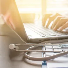 Image of a person sitting at a desk with hands on a lap top and a stethoscope and file folder nearby.