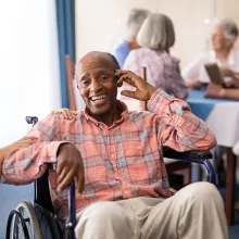 Female healthcare staff kneeling next to senior male in a wheelchair smiling at the camera in a healthcare facility setting.