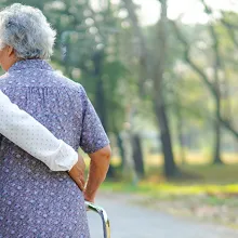 Female caregiver has arm around elderly woman with a walker walking outside.