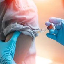 Image of hands in blue medical gloves administering a vaccine into the arm of a patient. The patient's shirt sleeve is rolled up.