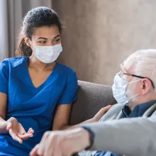 Doctor or female caregiver with senior man grandfather wearing protective mask to protect from Covid 19 at home or nursing home. Doctor talking consulting elderly patient at home