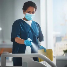 Image of health care worker wearing a mask and gloves and spraying a bed with a spray bottle