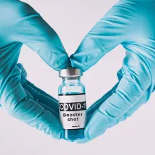 Gloved clinician making a heart with hands holding a vaccine vile 