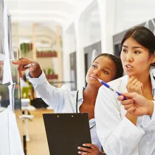 Image of a diverse group of doctors grouped together on the right pointing to and looking at a clear dry erase board on the left.