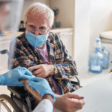 Image of older man wearing a medical mask and sitting in a wheel chair being attended to by a medical professional wearing am medical mask.