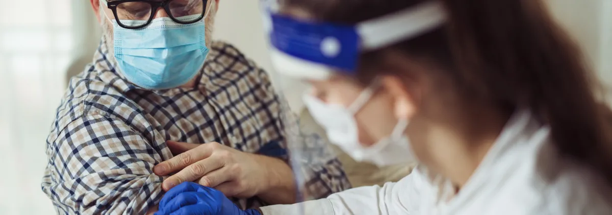 Young woman nurse with surgical mask and face shield giving injection to senior man at home or in a nursing home. 