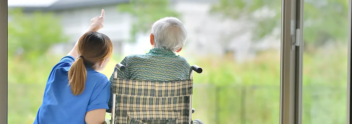 Image of nursing home staff kneeling next to man in wheelchair looking out the window.