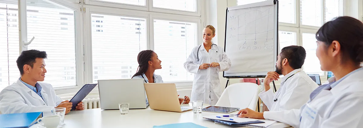 Healthcare providers in white coats sitting around table listening to doctor present.