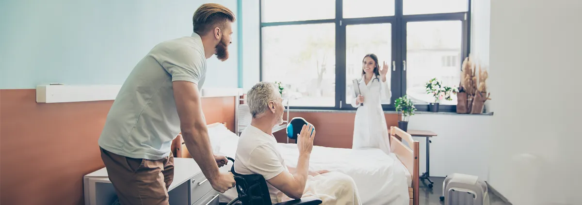 Male pushing elderly man in wheelchair out of room waving to clinician