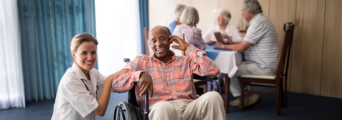 Female nurse in white jacket kneeling down next to male patient sitting in a wheelchair 