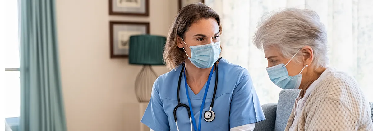 Female nurse with mask on sitting down with female elderly patient with mask on