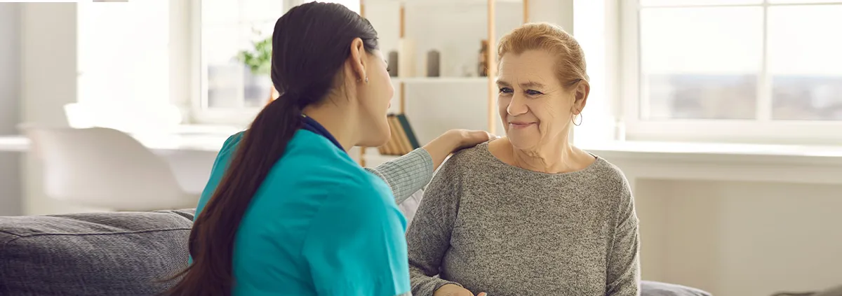 Friendly doctor supporting senior woman. Happy young nurse or clinician and her older adult patient sitting on sofa, looking at each other, holding hands, talking and smiling. 