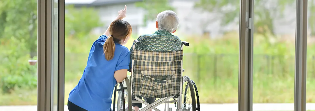 Image showing a health care worker kneeling beside an older man in a wheelchair as they look out two French doors.