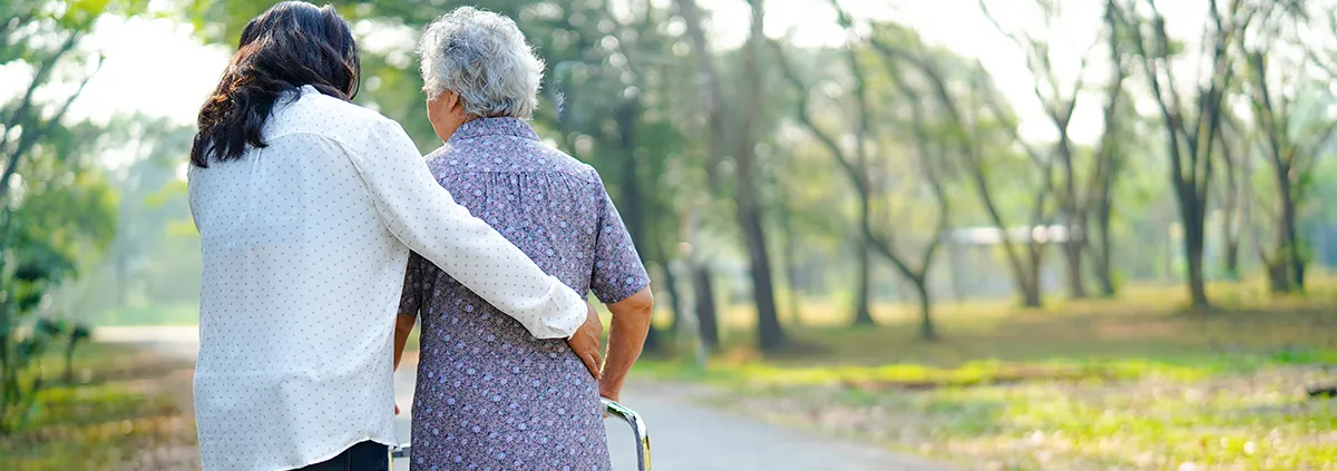 Female caregiver has arm around elderly woman with a walker walking outside.